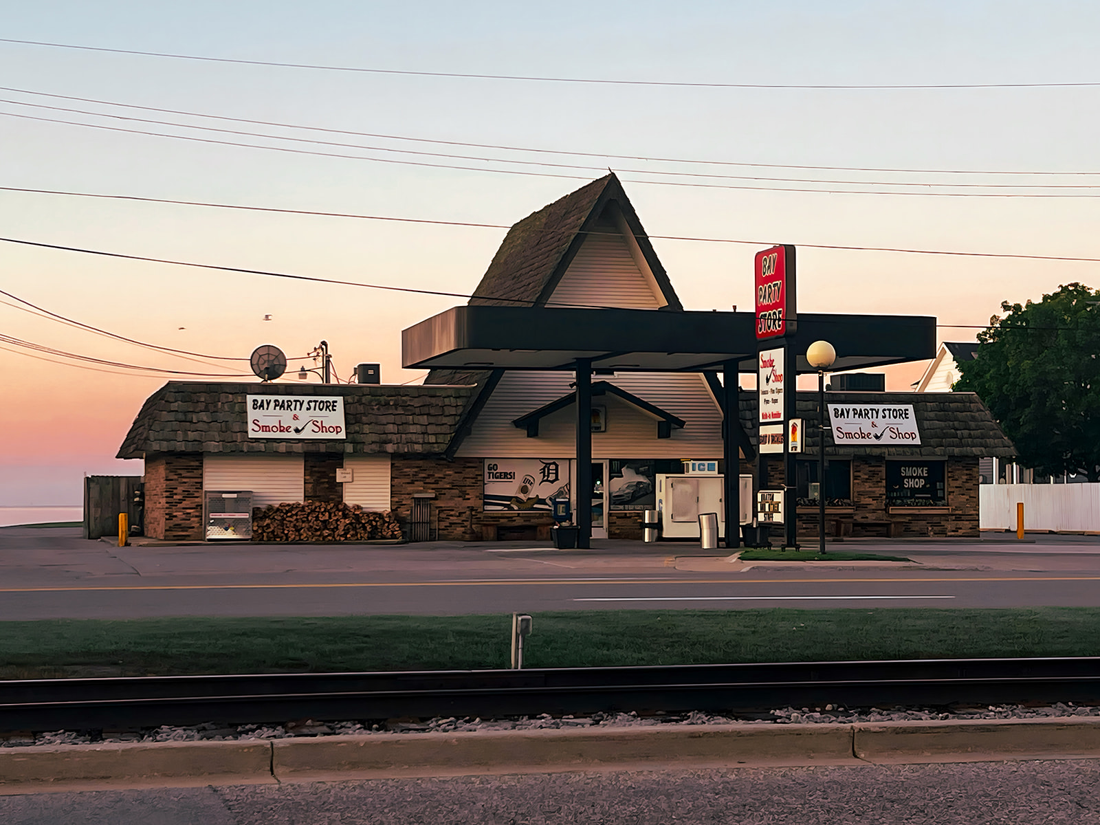 Bay Party Store & Smoke Shop — A-frame storefront at sunrise across the railroad tracks from the lake